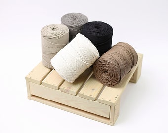 Puede incluir: Cinco carretes de cuerda de algodón en varios colores, incluyendo natural, beige, marrón, gris y negro, están dispuestos en una tarima de madera.
