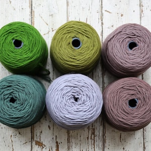 May include: Six skeins of yarn in various colors: green, olive green, teal, lavender, and two shades of mauve. The yarn is tightly wound into round balls, each with a central hole. The skeins are arranged on a distressed white wooden surface.