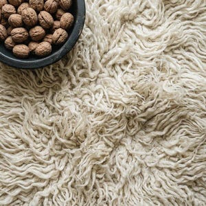 May include: A close-up of a shaggy, off-white rug with a textured, looped pile. A dark gray bowl filled with walnuts sits in the upper left corner. The rug's fibers create a soft, inviting appearance.