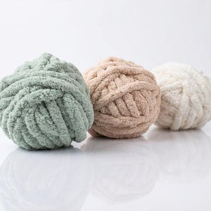 May include: Three balls of chunky yarn in a row, each with a different color. The colors are a soft green, a light tan, and a creamy white. The yarn is thick and textured, perfect for knitting or crocheting projects.