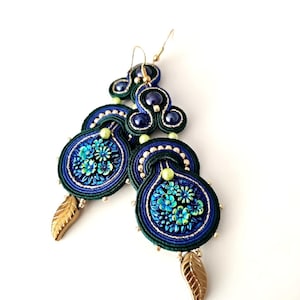 Puede incluir: Pendientes azules y verdes con detalles dorados y un dije de hoja. Los pendientes están hechos de hilo tejido y presentan un diseño floral.