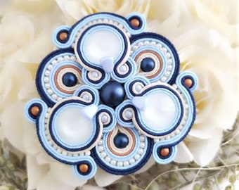 Puede incluir: Un broche de flor de cuentas azules, blancas y marrones con un centro nacarado. El broche está sobre un fondo blanco.