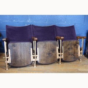 Puede incluir: Una butaca de cine vintage de tres plazas con respaldos y asientos de terciopelo morado. Los respaldos de madera son abatibles. Los reposabrazos son de madera y los soportes metálicos tienen un acabado de latón.