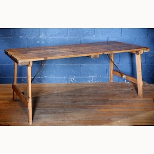 Puede incluir: Una mesa rectangular de madera con un acabado marrón desgastado y una superficie superior lisa. La mesa cuenta con cuatro patas robustas conectadas por soportes metálicos diagonales en cada lado, proporcionando estabilidad al armazón de madera.