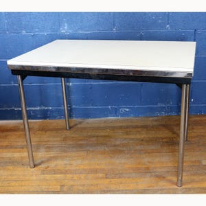 May include: A rectangular white table with a chrome trim and legs. The table is approximately 48 inches long, 30 inches wide, and 29 inches tall. The table has a black frame and is set against a blue wall and wooden floor.