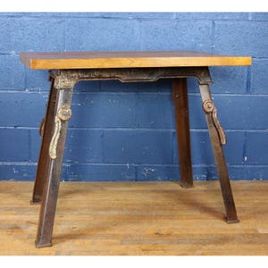 May include: A small, industrial-style table with a rectangular wooden top and a metal base. The base has a dark, aged finish with adjustable legs. The table is suitable for various uses, such as a side table or display stand.