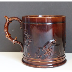 May include: A brown ceramic mug with a detailed, raised design of a person running. The mug has a handle shaped like a branch with leaves.