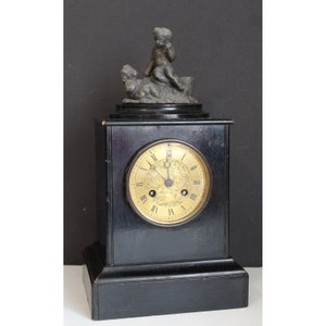 May include: A black wooden mantel clock with a gold face and Roman numeral hour markers. The clock is topped with a bronze sculpture of two cherubs.