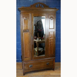 Può includere: Un alto armadio in legno marrone con un'anta centrale a specchio, due pannelli laterali con intagli circolari e un ampio cassetto alla base. La parte superiore dell'armadio è rifinita con una modanatura decorativa ad arco.