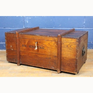 Op de afbeelding: Een vintage houten kist met een sleutelslot en metalen hardware. De kist heeft een warme bruine kleur met zichtbare houtnerf en de tekst "B.S. KEEPING" op de voorkant. Het heeft een rechthoekige vorm.