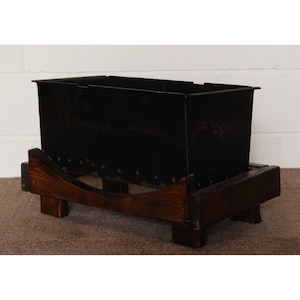 May include: A black rectangular metal planter box on a dark brown wooden base. The planter has a textured surface and is supported by a wooden frame with curved supports and four short legs. The design is rustic and traditional.