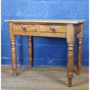 Puede incluir: Una pequeña mesa de madera antigua con dos cajones y patas torneadas. La mesa es de madera clara con un acabado natural. Los cajones tienen pomos redondos de madera. La mesa mide aproximadamente 76 cm de largo, 46 cm de ancho y 76 cm de alto.