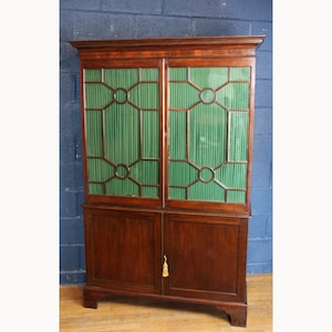 Op de afbeelding: Een hoge houten kast met twee glazen deuren boven twee massieve deuren. De glazen panelen hebben een groen, verticaal gestreept ontwerp binnen een geometrisch kader. Een decoratieve kwast hangt aan de onderste deuren.