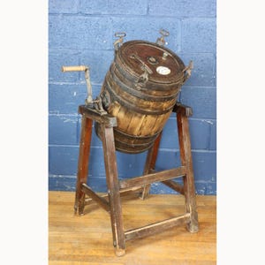 Puede incluir: Una antigua mantequera de madera con cuerpo en forma de barril, herrajes metálicos y manivela. La mantequera está montada sobre un soporte de madera con patas anguladas. La madera tiene un acabado natural, mostrando signos de edad y uso.