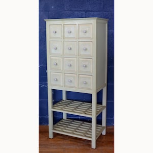 May include: A tall, off-white cabinet with sixteen small drawers and two open shelves. Each drawer has a small, round knob. The cabinet is painted in a light, neutral color and stands on a sturdy base. The cabinet is suitable for storage.