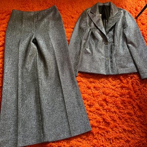 May include: A silver glitter two-piece suit, consisting of wide-leg pants and a blazer with a two-button closure. The suit is displayed on an orange textured surface. The blazer has two front pockets and a notched lapel.