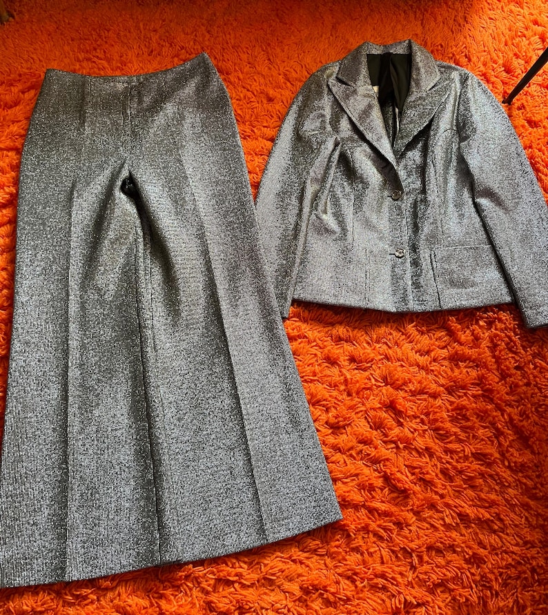 May include: A silver glitter two-piece suit, consisting of wide-leg pants and a blazer with a two-button closure. The suit is displayed on an orange textured surface. The blazer has two front pockets and a notched lapel.
