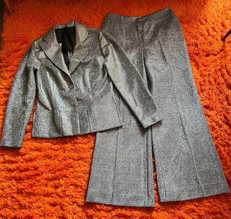 May include: A silver glitter suit set, including a blazer and wide-leg pants. The blazer has a notched collar and button closure. The pants have a high waist. The suit is displayed on an orange shag carpet.