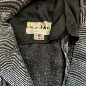May include: Close-up of a silver and black striped blazer. The blazer has a black lining and a label that reads "Peter Shires". The blazer is size 14.