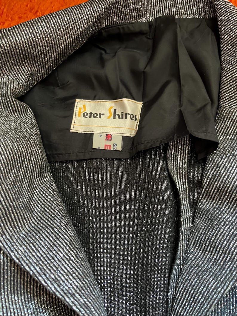 May include: Close-up of a silver and black striped blazer. The blazer has a black lining and a label that reads "Peter Shires". The blazer is size 14.