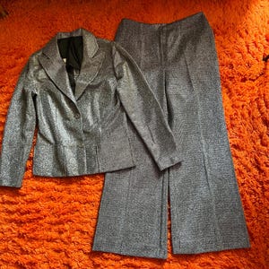 May include: A silver metallic blazer and matching wide-leg pants set. The blazer has a notched collar and button closure. The pants have a high waist and a flared leg. The fabric has a subtle shimmer, creating a glamorous look. The set is displayed on an orange surface.