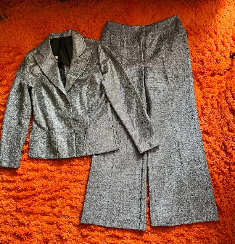 May include: A silver metallic blazer and matching wide-leg pants set. The blazer has a notched collar and button closure. The pants have a high waist and a flared leg. The fabric has a subtle shimmer, creating a glamorous look. The set is displayed on an orange surface.