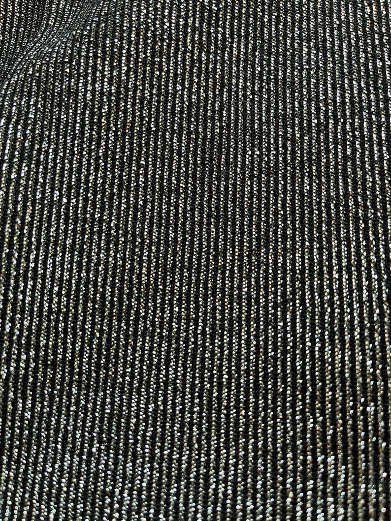 May include: Close-up of a black fabric with vertical silver metallic threads woven throughout. The threads create a shimmering, textured surface. The fabric has a subtle sheen, reflecting light and adding visual interest. The overall effect is a dark, elegant textile.
