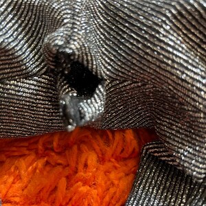 May include: Close-up of a silver and black fabric with a textured, wavy pattern. The fabric is folded, revealing a glimpse of bright orange, fluffy material underneath. The fabric has a metallic sheen.
