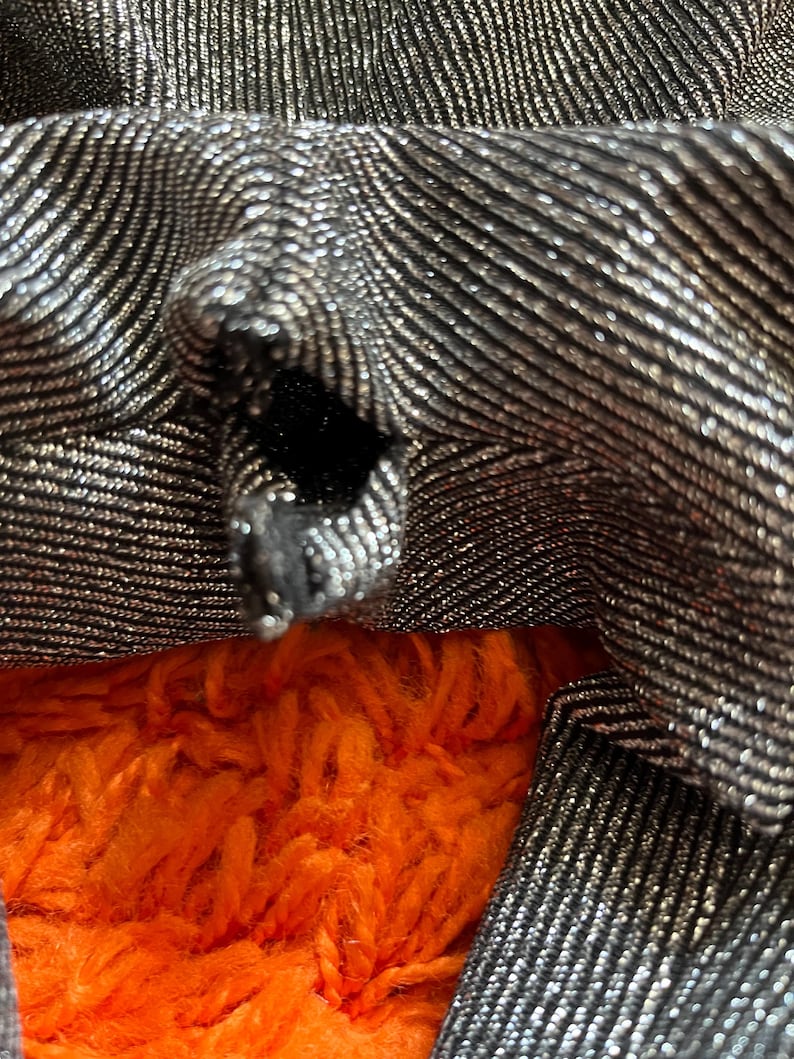 May include: Close-up of a silver and black fabric with a textured, wavy pattern. The fabric is folded, revealing a glimpse of bright orange, fluffy material underneath. The fabric has a metallic sheen.