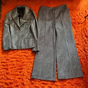 May include: A silver, shimmering two-piece suit set. The jacket has a notched collar and two buttons. The wide-leg pants are high-waisted. The suit is displayed on a bright orange, textured surface.
