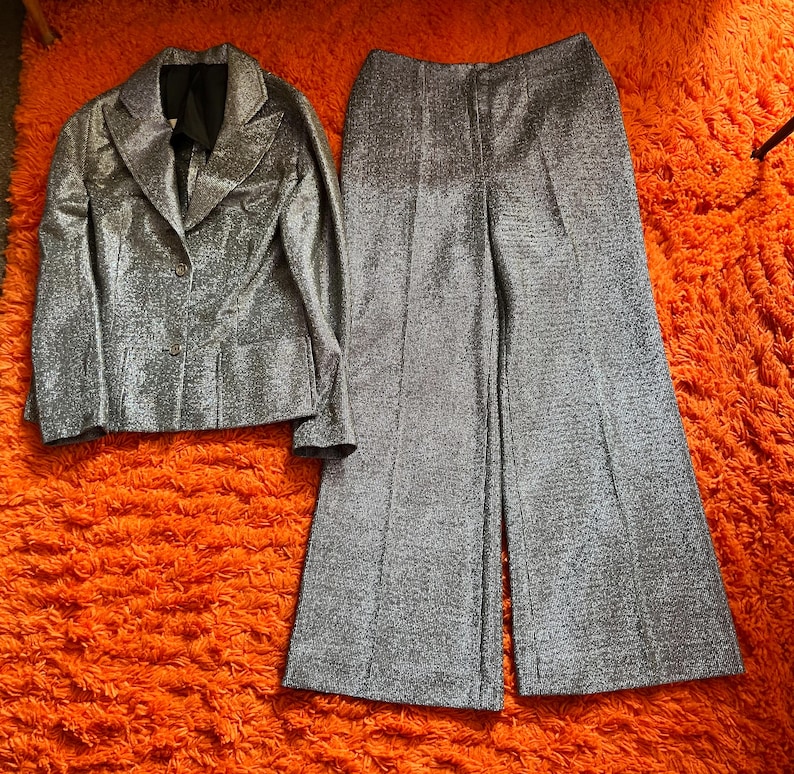 May include: A silver, shimmering two-piece suit set. The jacket has a notched collar and two buttons. The wide-leg pants are high-waisted. The suit is displayed on a bright orange, textured surface.