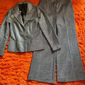 May include: A silver metallic two-piece suit, consisting of a blazer and wide-leg pants. The blazer has a notched collar and button closure. The pants have a high waist and a flared leg. The suit is displayed on an orange shag carpet.