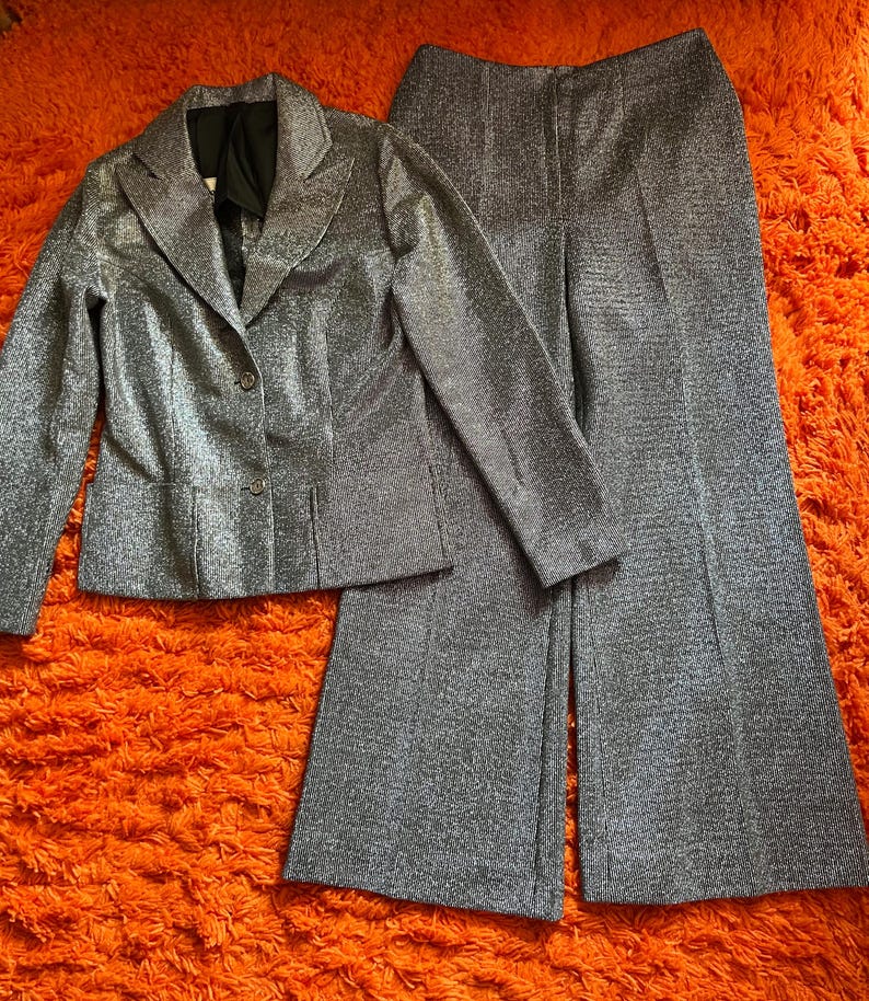 May include: A silver metallic two-piece suit, consisting of a blazer and wide-leg pants. The blazer has a notched collar and button closure. The pants have a high waist and a flared leg. The suit is displayed on an orange shag carpet.