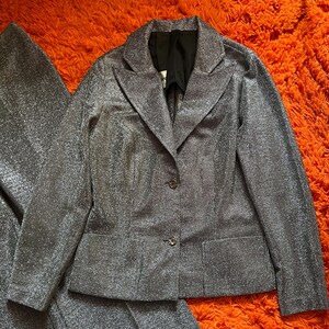 May include: A silver-colored suit set, including a blazer and matching pants, is displayed on an orange surface. The blazer has a notched collar, two buttons, and two front pockets. The fabric has a textured, shimmering appearance.
