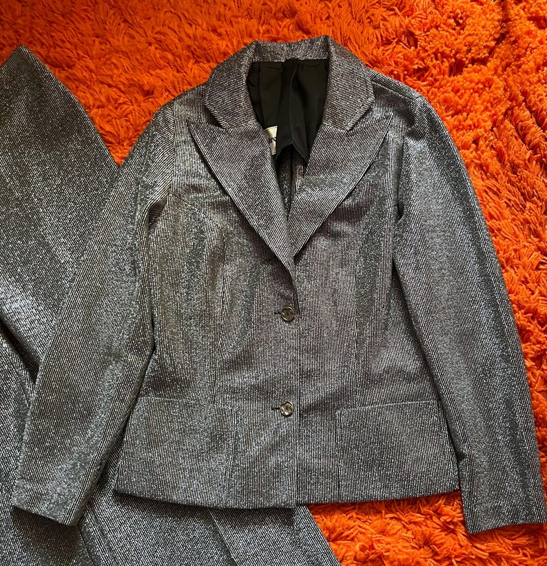May include: A silver-colored suit set, including a blazer and matching pants, is displayed on an orange surface. The blazer has a notched collar, two buttons, and two front pockets. The fabric has a textured, shimmering appearance.