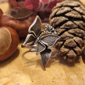 May include: A silver ring with a butterfly and leaf design. The ring features detailed textures on the butterfly wings and leaves. The ring is set against a natural background with a pine cone and conkers.
