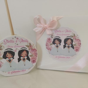 Può includere: Un diffusore in ceramica bianca e una scatola regalo bianca con un nastro rosa. Entrambi gli articoli presentano illustrazioni di due ragazze in abiti bianchi, con il testo "Stella e Perla Prima Comunione" e la data "22 Settembre 2024".