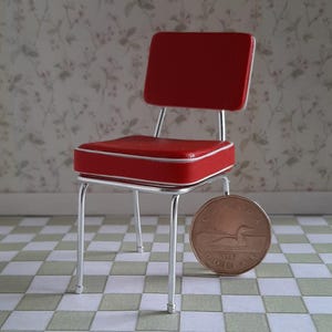 May include: A miniature red chair with a white piped edge and a silver metal frame. The chair is positioned next to a 1987 Canadian dollar coin, which serves as a scale reference to show the small size of this dollhouse-scale furniture piece.