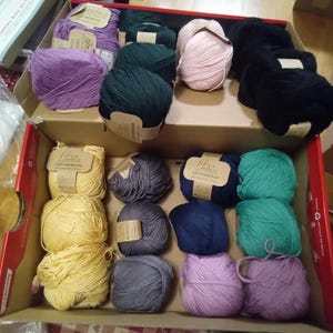May include: A variety of colorful yarn balls in a cardboard box. The yarn colors include purple, green, pink, yellow, gray, blue, and black. Each ball has a label attached. The box is red and brown.