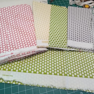 May include: Five fabric swatches with a polka dot pattern. The swatches are in pink, yellow, green, gray, and green. The swatches are all 100% organic cotton. The text "Birch Fabrics" and "modbasics" is visible on the pink swatch. The text "100% Organic Cotton" is visible on the green swatch.