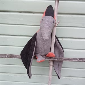 May include: A plush pterodactyl toy with gray, black, and orange accents. The toy is perched on a bamboo structure against a light green background. The pterodactyl has a long beak and large wings.