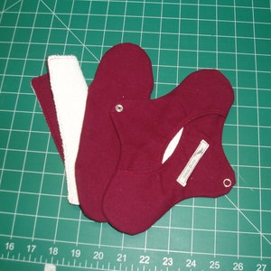 May include: Three fabric pieces for sewing a reusable menstrual pad. Two are burgundy and one is white. The burgundy pieces have a snap button and a small white label that says "Happy Days".