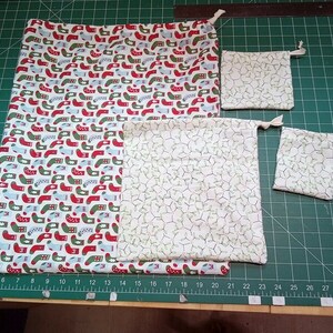 May include: Four fabric drawstring bags in various sizes. The largest bag is white with a red and green pattern of stockings. The other three bags are white with a green and white pattern of holly leaves.