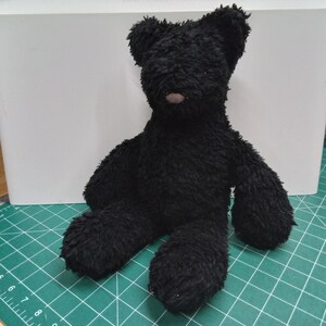 May include: A black teddy bear with a fluffy texture, sitting upright. The bear has a small, brown nose and is positioned on a green cutting mat. The background features a white surface.