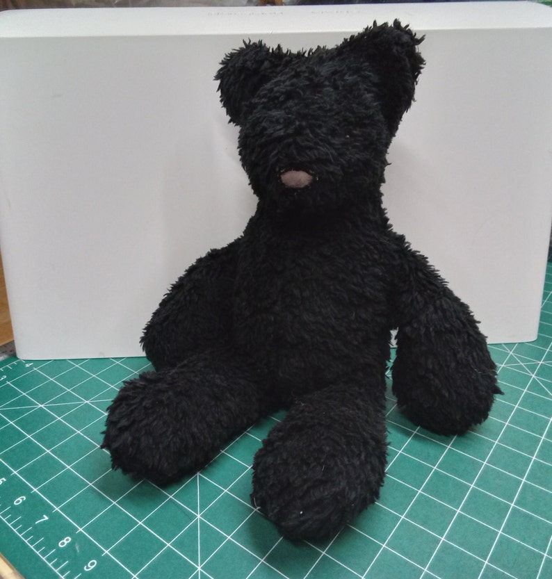 May include: A black teddy bear with a fluffy texture, sitting upright. The bear has a small, brown nose and is positioned on a green cutting mat. The background features a white surface.
