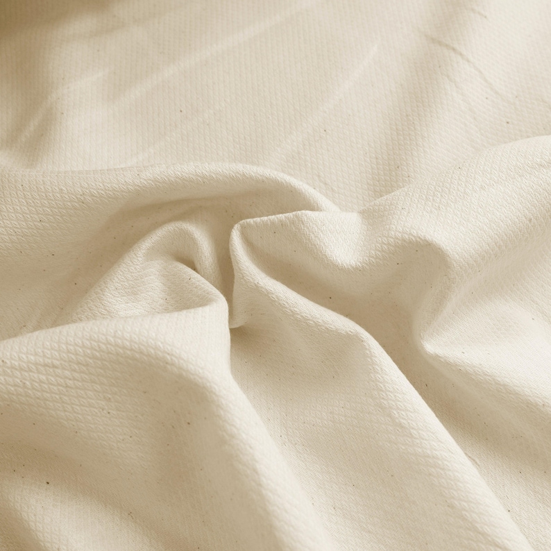 May include: Close-up of a textured, off-white fabric with a subtle woven pattern. The fabric is soft and drapes nicely.