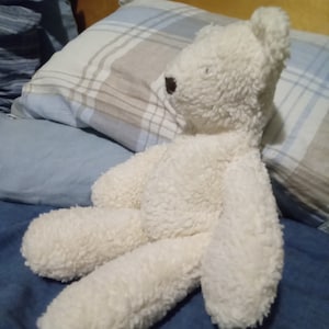 May include: A fluffy, off-white teddy bear with a brown nose and eyes. The bear is lying on a blue bedspread with a plaid pillow in the background. The bear's fur appears soft and textured.