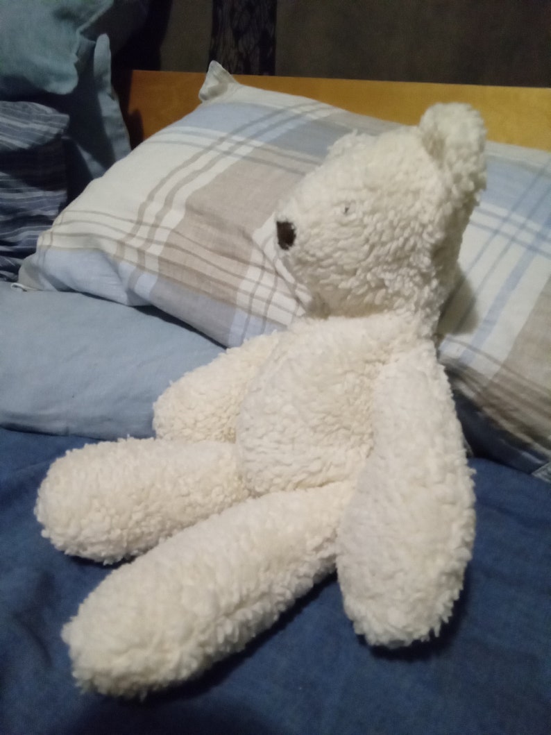 May include: A fluffy, off-white teddy bear with a brown nose and eyes. The bear is lying on a blue bedspread with a plaid pillow in the background. The bear's fur appears soft and textured.