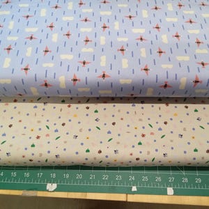 May include: Two rolls of patterned fabric. The top fabric is light blue with a repeating pattern of pink airplanes, white clouds, and vertical lines. The bottom fabric is off-white with a floral and dot pattern. A green cutting mat with a ruler is at the bottom.
