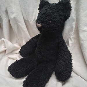 May include: A handmade black teddy bear with a soft, textured appearance. The bear has a light pink nose and small, round eyes. Its arms and legs are extended, and it is sitting on a white surface.